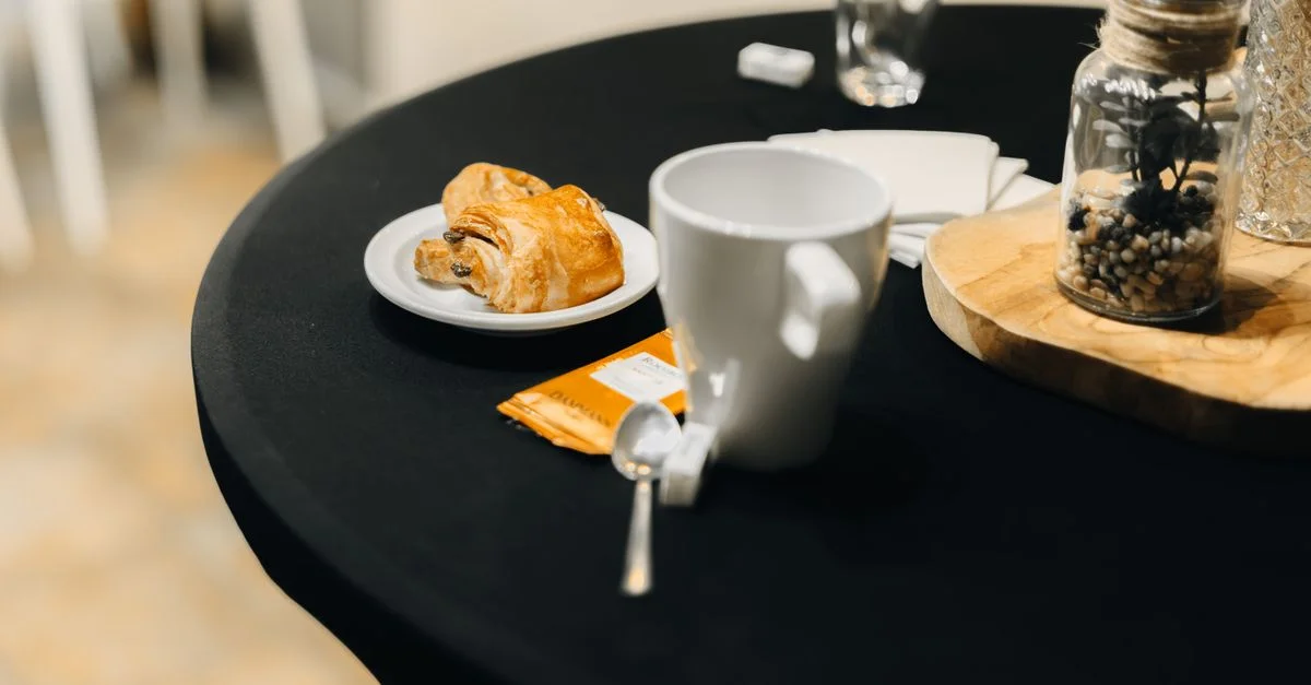 Un cadre de café confortable avec un croissant, une tasse et un bocal décoratif sur une table ronde. Idéal pour un brunch relaxant.